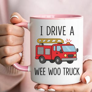 May include: White ceramic mug with a pink interior and handle. The mug features a red fire truck graphic and the text "I DRIVE A WEE WOO TRUCK" in black. The mug is held by a person.
