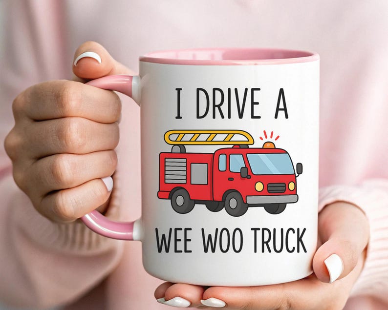May include: White ceramic mug with a pink interior and handle. The mug features a red fire truck graphic and the text "I DRIVE A WEE WOO TRUCK" in black. The mug is held by a person.