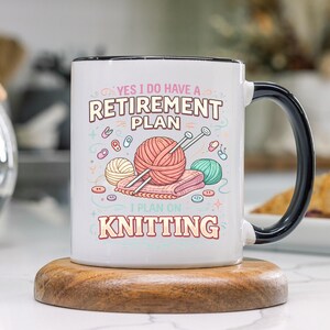 Op de afbeelding: Witte keramische mok met een zwarte handgreep en rand, met de tekst "YES I DO HAVE A RETIREMENT PLAN I PLAN ON KNITTING". Het ontwerp bevat illustraties van garen, breinaalden en knopen. De mok staat op een houten onderzetter.
