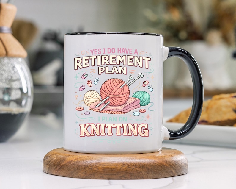 Op de afbeelding: Witte keramische mok met een zwarte handgreep en rand, met de tekst "YES I DO HAVE A RETIREMENT PLAN I PLAN ON KNITTING". Het ontwerp bevat illustraties van garen, breinaalden en knopen. De mok staat op een houten onderzetter.