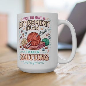 Op de afbeelding: Witte keramische mok met de tekst "YES I DO HAVE A RETIREMENT PLAN I PLAN ON KNITTING". De mok heeft illustraties van garen, breinaalden en knopen. De mok staat op een houten oppervlak.