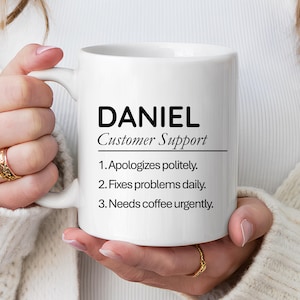 Può includere: Tazza in ceramica bianca con il nome "DANIEL" e il titolo "Customer Support" stampati in nero. La tazza elenca tre affermazioni: "Si scusa gentilmente", "Risolve i problemi quotidianamente" e "Ha urgentemente bisogno di caffè."