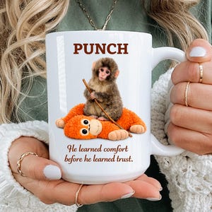 Könnte beinhalten: Weiße Keramiktasse mit einem Cartoon-Affen-Design. Das Wort "PUNCH" ist in Braun über dem Affen gedruckt, der einen Stock hält und auf einem orangefarbenen Plüschtier sitzt. Der Text "He learned comfort before he learned trust." steht darunter.
