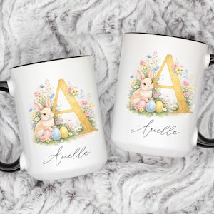 Pode incluir: Duas canecas de cerâmica branca com alças e bordas pretas. Cada caneca apresenta um desenho em aquarela de um coelho, ovos de Páscoa e uma letra "A" dourada rodeada por flores. O nome "Arielle" está escrito abaixo.