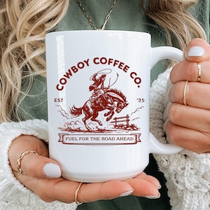 以下が含まれることがあります： 白いセラミックコーヒーマグ、ハンドル付き、茶色のグラフィックで馬に乗ったカウボーイ、"Cowboy Coffee Co."と"Fuel for the Road Ahead"の文字。マグカップは人が持っています。