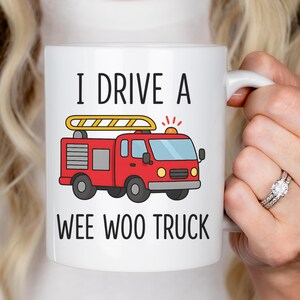 May include: White ceramic mug with the text "I DRIVE A WEE WOO TRUCK" and a cartoon illustration of a red fire truck. The fire truck has a yellow ladder and flashing lights.