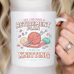 Op de afbeelding: Witte keramische mok met de tekst "YES I DO HAVE A RETIREMENT PLAN I PLAN ON KNITTING" en een kleurrijk brei-ontwerp. Het ontwerp bevat bollen wol, breinaalden en knopen.
