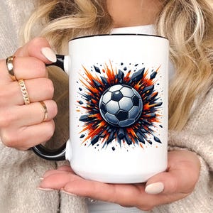May include: White ceramic mug with a black handle and rim. The mug features a soccer ball design at the center, surrounded by an abstract explosion of orange, blue, and black shapes. The mug is held by a person.