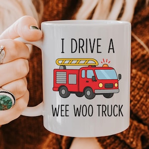 May include: White ceramic mug with the text "I DRIVE A WEE WOO TRUCK" and a cartoon illustration of a red fire truck with a yellow ladder. The mug is held by a person.