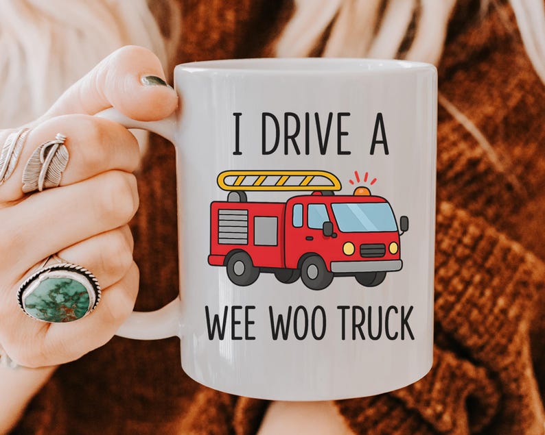 May include: White ceramic mug with the text "I DRIVE A WEE WOO TRUCK" and a cartoon illustration of a red fire truck with a yellow ladder. The mug is held by a person.