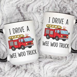 May include: Two white coffee mugs with black handles and rims. Each mug features a red fire truck graphic with the text "I DRIVE A WEE WOO TRUCK". The mugs are on a fluffy, white surface.