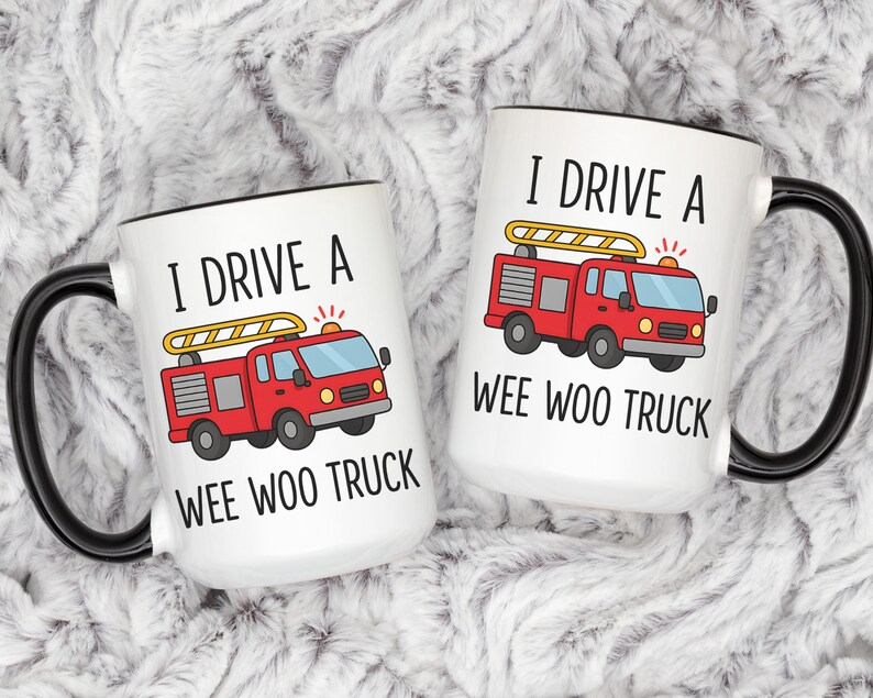 May include: Two white coffee mugs with black handles and rims. Each mug features a red fire truck graphic with the text "I DRIVE A WEE WOO TRUCK". The mugs are on a fluffy, white surface.