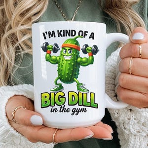 以下が含まれることがあります： 白いセラミックマグカップ。漫画のピクルスのキャラクターがダンベルを持ち上げています。ピクルスはヘッドバンドとスニーカーを着用し、テキスト「I'M KIND OF A BIG DILL in the gym」は緑と白です。