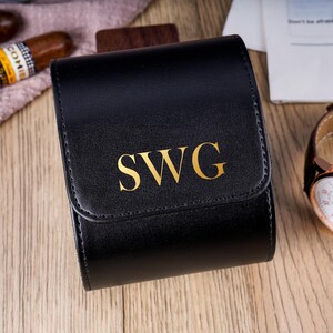 Puede incluir: Estuche de reloj de cuero negro con las iniciales doradas "SWG". Un reloj de cuero marrón con esfera blanca y detalles dorados está al lado. Dos puros sugieren un tema de lujo. El conjunto está sobre una superficie de madera.