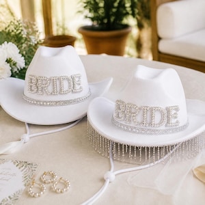 Könnte beinhalten: Zwei weiße Cowboyhüte mit dem Wort BRIDE in großen, silberfarbenen, mit Strasssteinen besetzten Buchstaben auf der Vorderseite. Jeder Hut hat ein passendes Strassband um die Krone, und ein Hut ist mit langen, herabhängenden Silberfransen versehen.