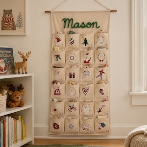 May include: A beige fabric wall-hanging advent calendar featuring the name Mason in green cursive script. It has twenty-four individual pockets, each embroidered with a unique red, green, and white holiday-themed icon and a corresponding number.