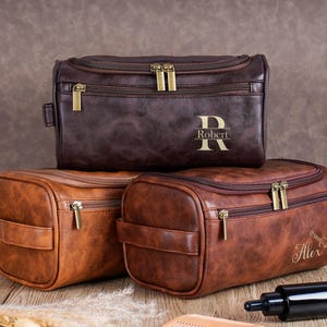 May include: Three brown leather toiletry bags with gold zippers and handles. The top bag is dark brown and has the name "Robert" printed on it. The bottom two bags are lighter brown and have the name "Alex" printed on one.