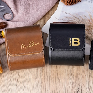 Könnte beinhalten: Zwei Leder-Uhrenetuis, eines braun mit "Maddex" in goldener Schrift, das andere schwarz mit "Bennett" und einem goldenen "B". Eine Uhr mit schwarzem Armband und ein Zigarrenschneider sind ebenfalls sichtbar.