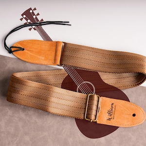 Puede incluir: Una correa de guitarra marrón con un diseño tejido y extremos de cuero. La correa tiene una hebilla de metal y el texto "William BEST GUITARIST" está impreso en el extremo de cuero. Una ilustración de guitarra está en el fondo.