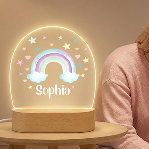 May include: A wooden-based night light with a rainbow design and the name "Sophia" etched into the acrylic. The rainbow features pastel colors, and the light is on a small, round wooden table.