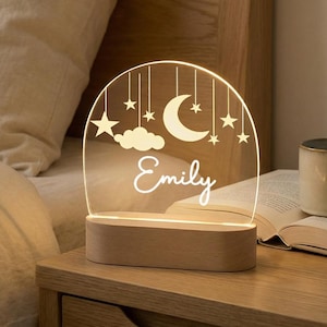 May include: A personalized night light with a wooden base and a clear acrylic top. The acrylic features a crescent moon, stars, and clouds design, with the name "Emily" in a script font. The lamp is on a wooden nightstand.