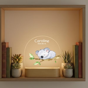 Könnte beinhalten: Ein Holzregal zeigt ein dekoratives Nachtlicht mit einer Illustration eines schlafenden Koalas. Das Licht ist mit dem Namen "Caroline" und dem Datum "2024.06.15" personalisiert. Das Regal enthält auch kleine Topfpflanzen und Bücher.