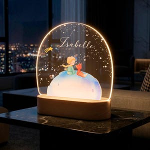 May include: A decorative night light featuring a whimsical illustration of a character with a fox on a planet, with the name "Isabelle" in script. The light has a wooden base and a clear, arched top with a warm glow, perfect for a child's room.