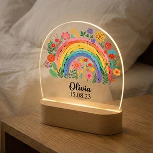 May include: A personalized night light featuring a colorful rainbow design with floral accents. The clear acrylic panel is set on a wooden base, with the name "Olivia" and date "15.08.23" printed below the rainbow. The light is on, illuminating the design.