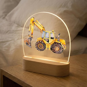 Puede incluir: Una lámpara de noche con forma de excavadora amarilla con diseño de acuarela. La excavadora está sobre un panel acrílico transparente arqueado, iluminado por un brillo cálido. La base es de madera clara, creando un ambiente suave y acogedor.