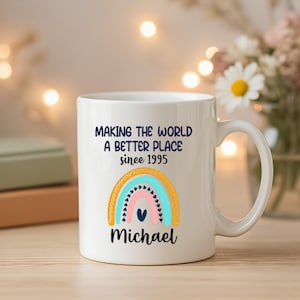 May include: White ceramic mug with a rainbow design and the text "MAKING THE WORLD A BETTER PLACE since 1995" and the name "Michael". The rainbow features blue, pink, and yellow colors.