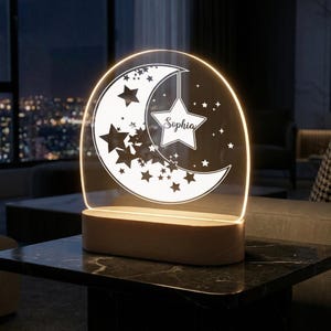 May include: A personalized night light featuring a crescent moon and stars design. The clear acrylic panel is illuminated by warm light, with the name "Sophia" displayed on a star. The base is made of wood.
