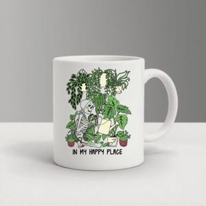 Puede incluir: Taza de cerámica blanca con una ilustración verde y blanca de un esqueleto cuidando plantas. El texto "IN MY HAPPY PLACE" está impreso debajo. La taza tiene un asa curvada.