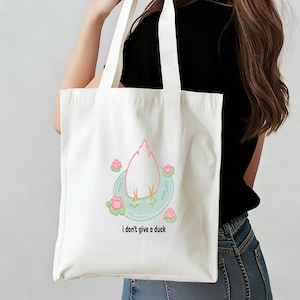 May include: A white tote bag featuring a cartoon duck with its rear end up in a pond with pink water lilies. The text "i don't give a duck" is printed below the duck illustration. The bag has two white handles.