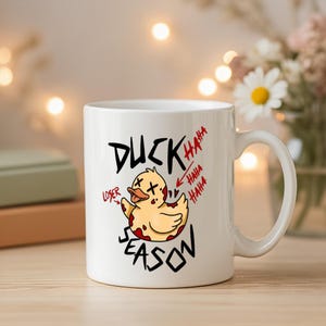 May include: White ceramic mug with a cartoon duck graphic. The duck is yellow with red accents and the text "DUCK SEASON" and "LOSER" are printed on the mug. The mug also has the text "HAHA" in red.