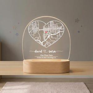 Op de afbeelding: Een gepersonaliseerd nachtlampje met een hartvormig kaartontwerp. Het heldere acryl paneel toont een stadskaart met een rood hart dat een locatie markeert. De basis is gemaakt van lichtgekleurd hout, en de tekst luidt "david & sara, Our First Date, October 26 2023 | San Diego, CA."