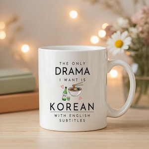 May include: White ceramic mug with the text "THE ONLY DRAMA I WANT IS KOREAN WITH ENGLISH SUBTITLES." The mug features a graphic of a bowl of noodles, a bottle of soju, and a small cup. The mug has a curved handle.