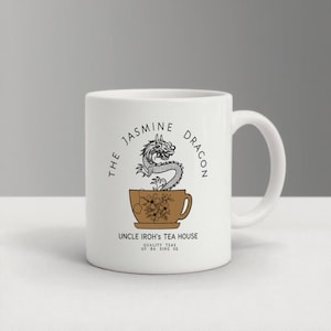 Op de afbeelding: Witte keramische mok met een drakenontwerp en de tekst "The Jasmine Dragon" en "Uncle Iroh's Tea House". De mok heeft een bruine theekopjesafbeelding met bloemaccenten en de woorden "Quality Teas of Ba Sing Se".