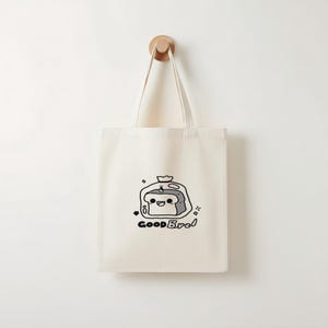 May include: Cream-colored tote bag with a cartoon illustration of a slice of bread in a plastic bag. The bread has a smiling face and the words "Good Bred" are written below the image in black. The bag hangs from a wooden hook.