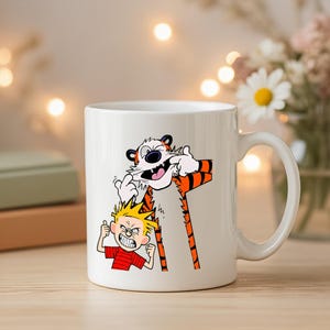 May include: White ceramic mug featuring a cartoon illustration of Calvin and Hobbes. The design shows Calvin with a tiger, both making silly faces. The mug has a curved handle and is suitable for hot or cold beverages.
