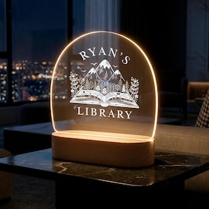 May include: A personalized night light with the text "Ryan's Library" and a mountain and book design. The light has a clear acrylic panel with a warm glow and a wooden base. The lamp is on a black table.
