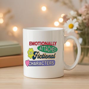 May include: White ceramic mug with the text "Emotionally Attached to Fictional Characters" in colorful lettering. The mug has a curved handle and is set against a blurred background with soft lighting and flowers.
