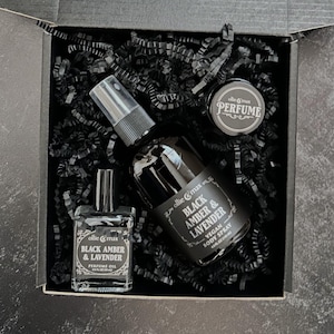 May include: A black gift box contains a perfume oil bottle, a body spray bottle, and a small jar of perfume. The bottles and jar are labeled with the text "Black Amber & Lavender" and the brand name "ollie & max". The box is filled with black paper.