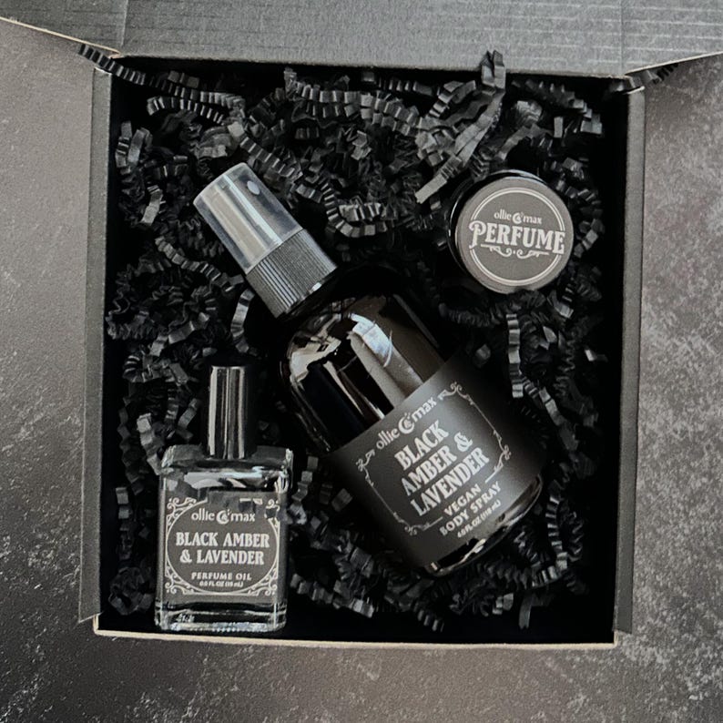 May include: A black gift box contains a perfume oil bottle, a body spray bottle, and a small jar of perfume. The bottles and jar are labeled with the text "Black Amber & Lavender" and the brand name "ollie & max". The box is filled with black paper.