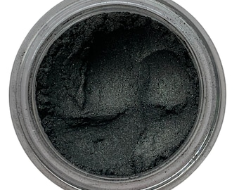 May include: A jar filled with dark green powder, possibly eyeshadow or pigment. The powder has a slightly shimmery appearance and is contained within a clear, round jar with a silver lid. The jar is set against a white background.