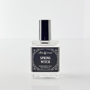 May include: A clear glass perfume oil bottle with a black cap. The label reads "ollie & max SPRING WITCH PERFUME OIL 0.5 FL OZ (15 mL)" in white text on a black background. The bottle is square and sits on a white surface.
