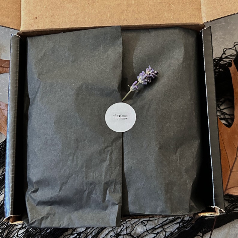 May include: An open cardboard box with black tissue paper inside. A sprig of lavender rests on top of the paper. A white sticker with the text "ollie & max" is on the tissue paper.