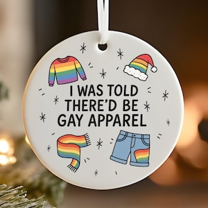May include: A white ceramic Christmas ornament with the text "I WAS TOLD THERE'D BE GAY APPAREL" and illustrations of a rainbow sweater, hat, scarf, and shorts. The ornament hangs from a white ribbon and is decorated with small star shapes.