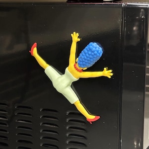May include: A magnet depicting a cartoon character with yellow skin, a blue beehive hairstyle, and a white dress. The character is in a dynamic pose with arms and legs outstretched, wearing red shoes and a red necklace. The magnet is attached to a black surface.