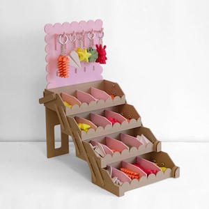 May include: A tiered brown display stand with scalloped edges and pink dividers, featuring a pink pegboard back panel. Five keychains in orange, white, yellow, green, and red are hanging from the top hooks, while small items fill the bins.