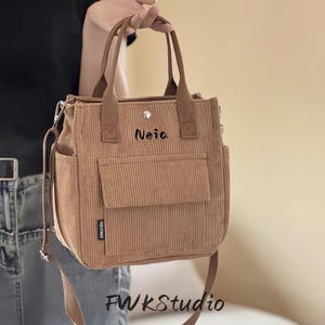Puede incluir: Bolso tote de pana marrón claro con un bolsillo frontal y una correa para el hombro. El bolso tiene el nombre "Neia" bordado en la parte delantera y el texto "FWKStudio" en la parte inferior.